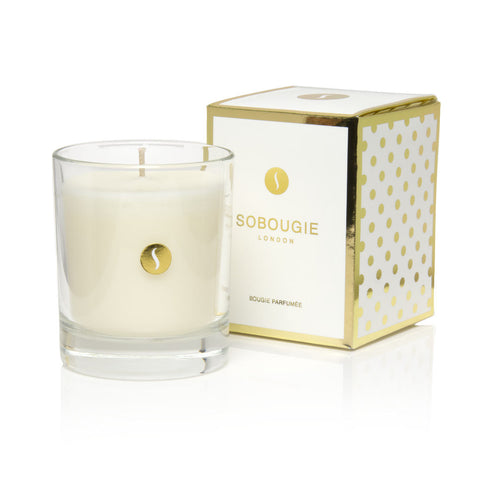 An unlit SOBOUGIE Blanc Velours luxury scented candle in a clear glass jar next to its signature white and metallic gold gift box packaging, containing an intimate fragrance profile of creamy musk, ambrox, fresh coconut water, and delicate rose.