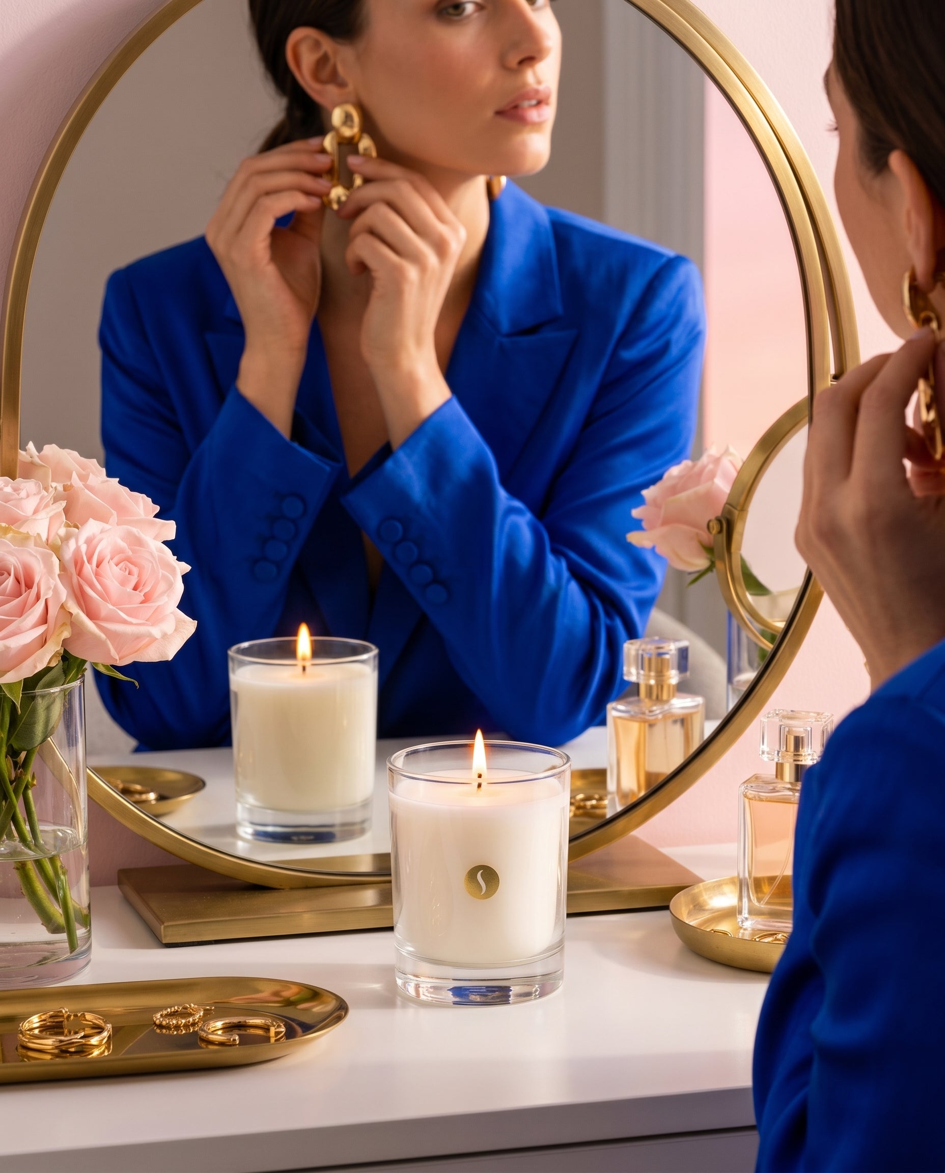 A lit SOBOUGIE luxury scented candle on a white vanity table with gold jewelry trays, pink roses, and perfume bottles, reflecting a woman in a royal blue blazer adjusting her gold earrings in a round brass mirror.