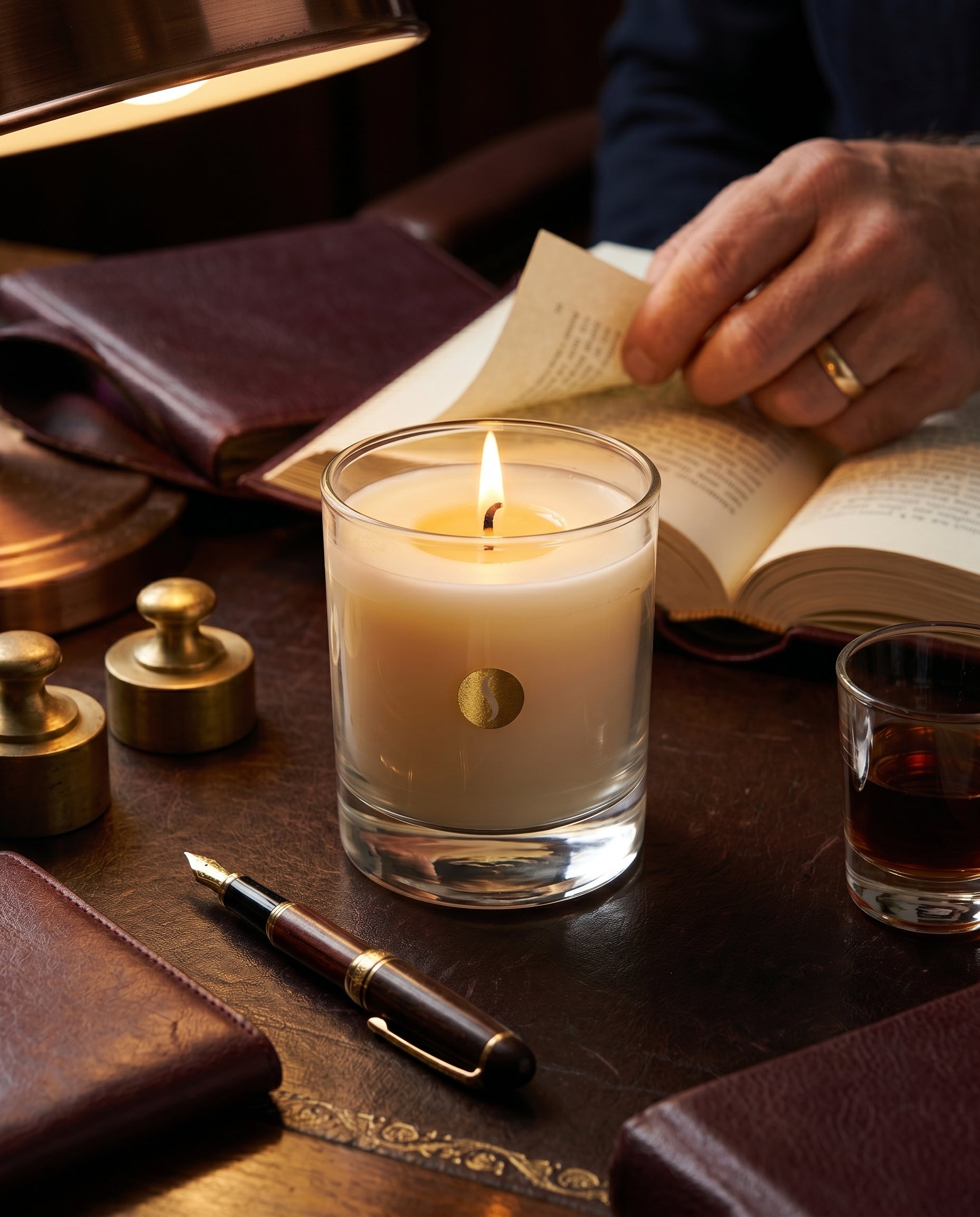 A lit SOBOUGIE luxury scented candle in a clear glass jar on a dark leather desk, surrounded by a fountain pen, leather notebooks, a glass of whiskey, and a man reading a book under warm lamp lighting.